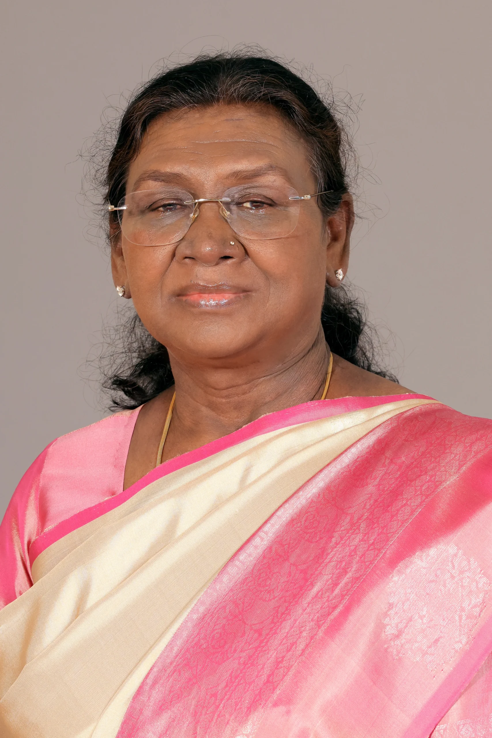 Official portrait of Droupadi Murmu, the 15th President of India, wearing a pink and cream silk saree and glasses against a neutral background.